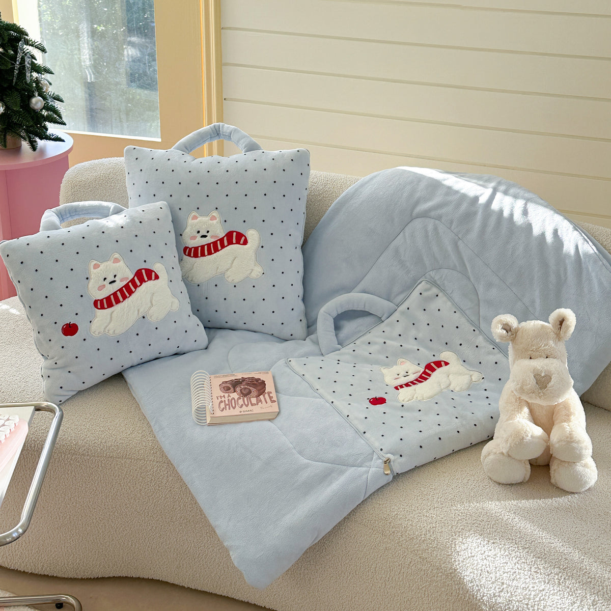 Puppy pillow blanket fully unfolded into a large baby blue polka dot throw, with two zipped cushion versions and a plush westie dog toy arranged on a cream sofa beside a small Christmas tree