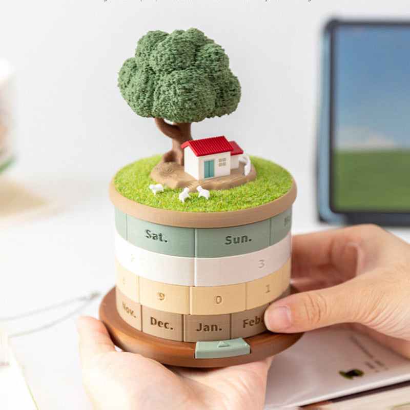 Two hands turning the month ring of the treehouse perpetual calendar - showing Nov Dec Jan Feb months, the grass meadow lid with cottage tree and sheep lifted slightly, rings for weekday and dates visible