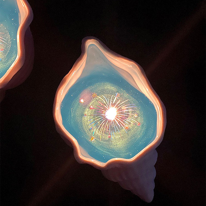 Close-up of a lit seashell candle against a dark background - pearlescent sculpted shell, translucent blue jelly wax shimmering with gold glitter, and a ring of holographic firework sparkles around the flame