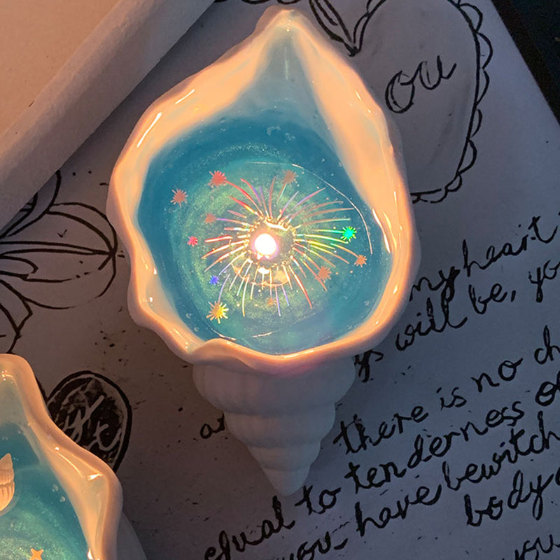 Two Fireworks at Sea shell candles glowing on a handwritten letter background - pearlescent conch shells with ocean-blue jelly wax and rainbow holographic firework sparkles around each cotton wick flame