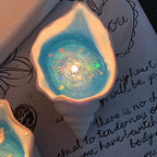 Two Fireworks at Sea shell candles glowing on a handwritten letter background - pearlescent conch shells with ocean-blue jelly wax and rainbow holographic firework sparkles around each cotton wick flame
