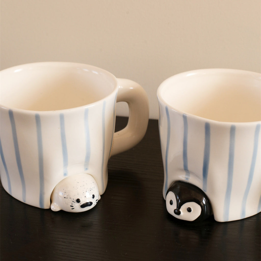 Close-up of the peekaboo seal and penguin mugs on a dark wood table - speckled cream seal and glossy black penguin sculpted peeking out from under hand-painted blue-striped ceramic bodies