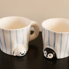 Close-up of the peekaboo seal and penguin mugs on a dark wood table - speckled cream seal and glossy black penguin sculpted peeking out from under hand-painted blue-striped ceramic bodies