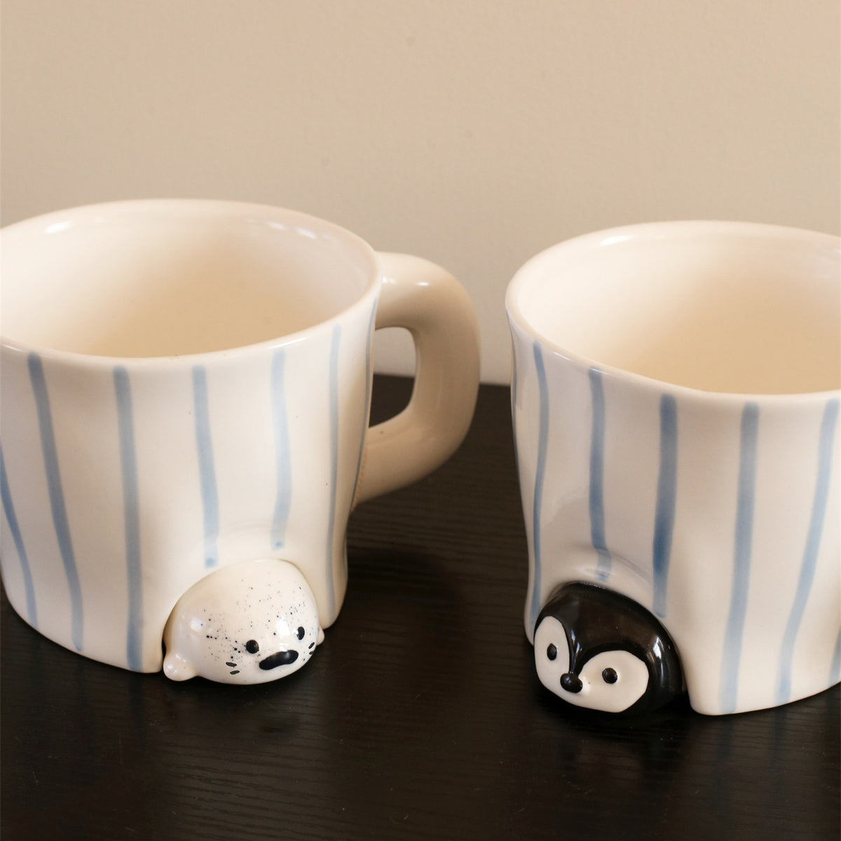 Close-up of the peekaboo seal and penguin mugs on a dark wood table - speckled cream seal and glossy black penguin sculpted peeking out from under hand-painted blue-striped ceramic bodies