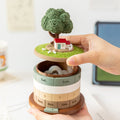 Hand lifting the grass meadow lid off the treehouse perpetual calendar to reveal the hidden storage compartment inside - rings showing Sat Sun, dates 6 7 and months Nov Dec Jan Feb, tablet in the background