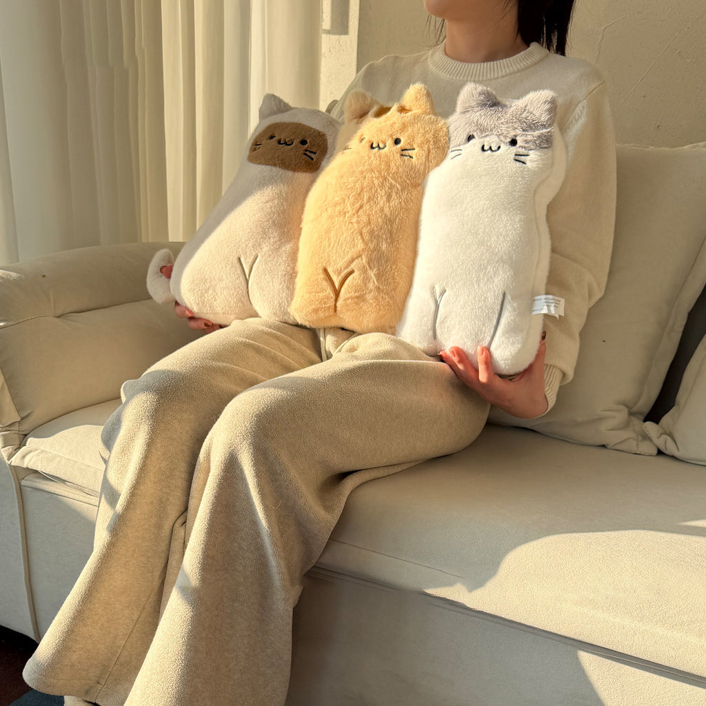 Woman in beige loungewear sitting on a sofa holding three long-body cat hug pillows - cow-cat, ginger tabby and grey-faced kitty plush cushions with embroidered faces