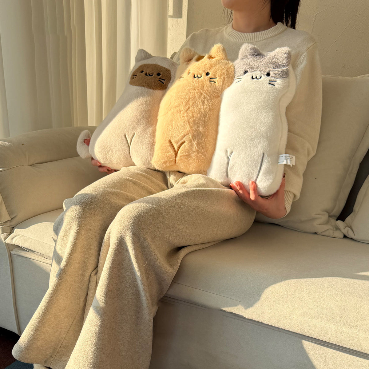 Woman in beige loungewear sitting on a sofa holding three long-body cat hug pillows - cow-cat, ginger tabby and grey-faced kitty plush cushions with embroidered faces