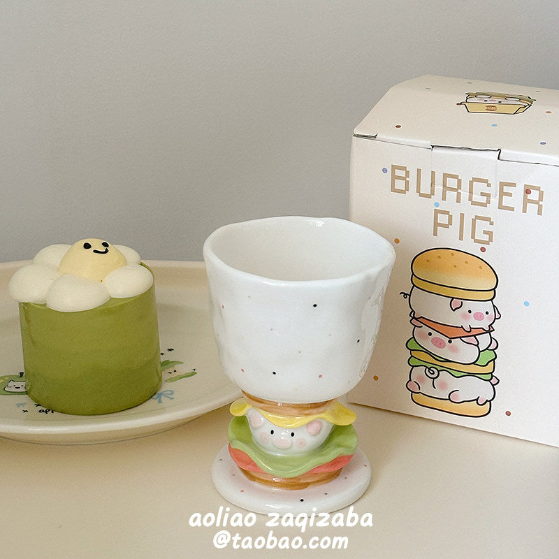 Burger Pig goblet mug on a cream table beside a matcha dessert and its dotted gift box - speckled white ceramic goblet on the sculpted burger stem with piggy face, lettuce and tomato layers, kawaii dessert scene