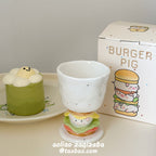 Burger Pig goblet mug on a cream table beside a matcha dessert and its dotted gift box - speckled white ceramic goblet on the sculpted burger stem with piggy face, lettuce and tomato layers, kawaii dessert scene