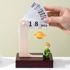 Hand flipping the month cards of the floating planet perpetual calendar - fanned cards from JAN to DEC visible above the frame, golden Saturn floating below with the reading prince figurine on the wooden base