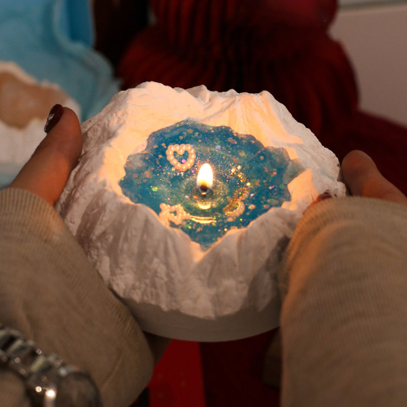 Two hands cupping the lit Glacier Ocean candle to show its size - snow-white plaster iceberg with a glowing blue jelly wax pool, pearl heart and bow charms, warm candlelight on a cozy background