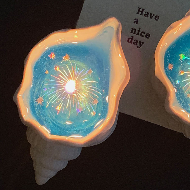 A lit Fireworks at Sea shell candle in the foreground with a second candle behind, next to a white Have a nice day card - blue jelly wax glowing with holographic firework sparkles inside a pearly sculpted shell