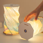 Hand unfolding the Starry Night folding lamp by pulling the top and base apart - mid-fold accordion paper shade with starry sky print catching warm light, a second fully lit lamp behind, octagonal top cap with power button visible