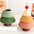 Hand dropping a pearl bracelet into the open green Christmas tree jewelry box - showing the hidden storage tier, with the cinnamon version fully stacked beside it on a white dressing table