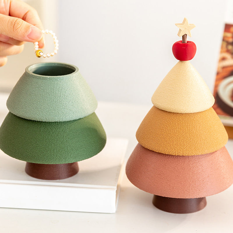 Hand dropping a pearl bracelet into the open green Christmas tree jewelry box - showing the hidden storage tier, with the cinnamon version fully stacked beside it on a white dressing table