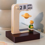 Reading prince figurine sitting with an open book and tea cup under the floating golden Saturn, cream flip calendar showing 28 APR WED, warm evening light on a desk corner