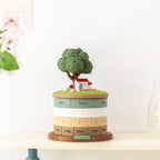 Treehouse perpetual calendar on a cream shelf against a white wall - miniature tree, cottage and three sheep on the grass lid, rings showing Sun Mon Tue, date 3, months Jan Feb Mar Apr, cherry blossom blurred at the side