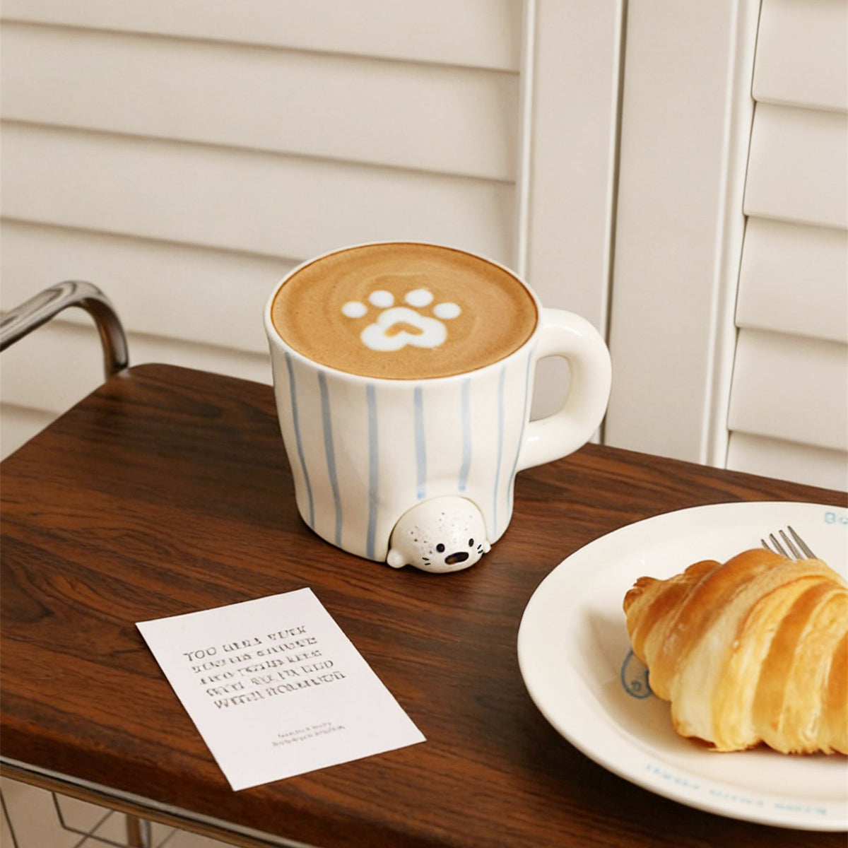 Peekaboo seal mug filled with paw-print latte art on a wooden cart - hand-painted blue striped ceramic cup with the speckled baby seal peeking out, a croissant on a white plate and a small poem card beside