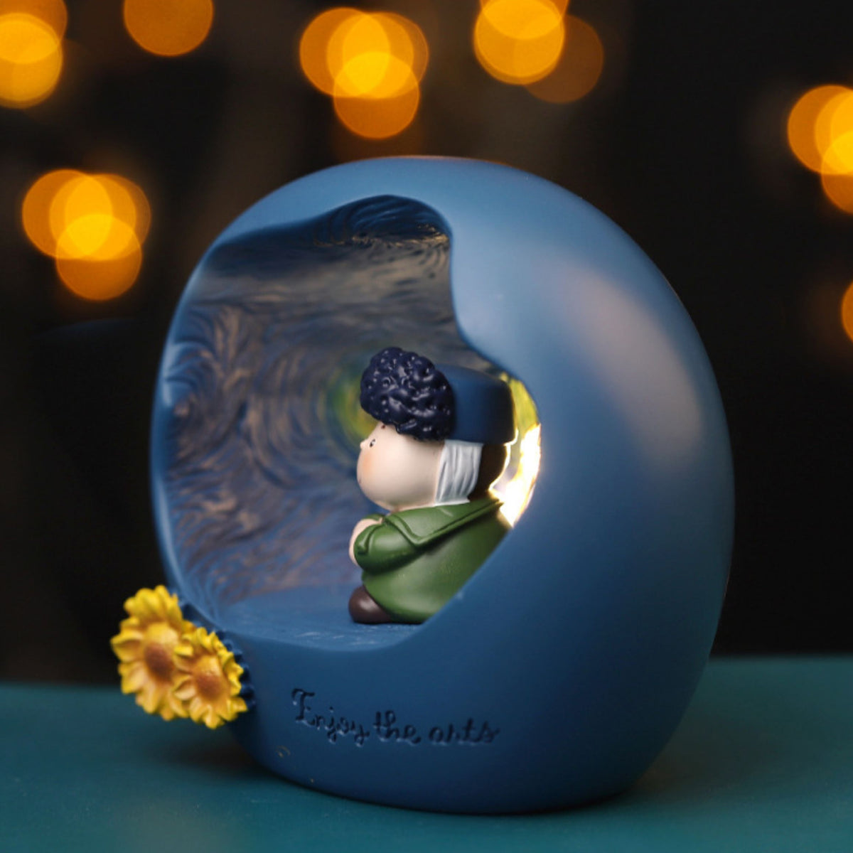 Side view of the Van Gogh art night light at night - the blue sphere glowing from within with bright warm light spilling through the carved opening, chibi painter figure in profile, sunflower and Enjoy the arts lettering visible, golden bokeh background