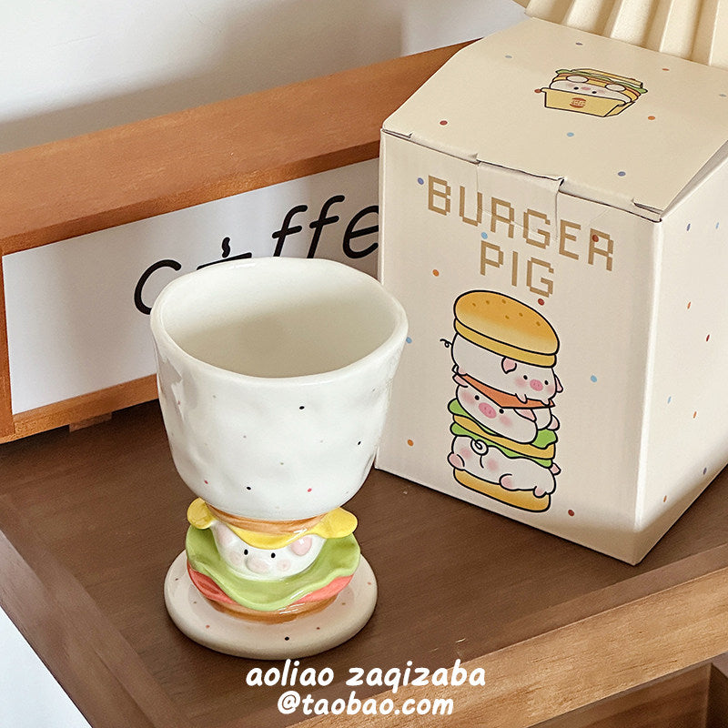 Burger Pig goblet mug on a wooden shelf next to its dotted gift box and a coffee sign - speckled white ceramic goblet on the 3D burger stem with piggy face tucked in the layers, warm cafe-style display