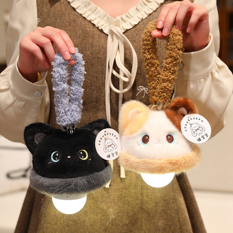 Two hands holding the lit black cat and calico cat plush night light charms by their fluffy loops, showing the moon-and-star eyes on the black cat and both warm bulbs glowing