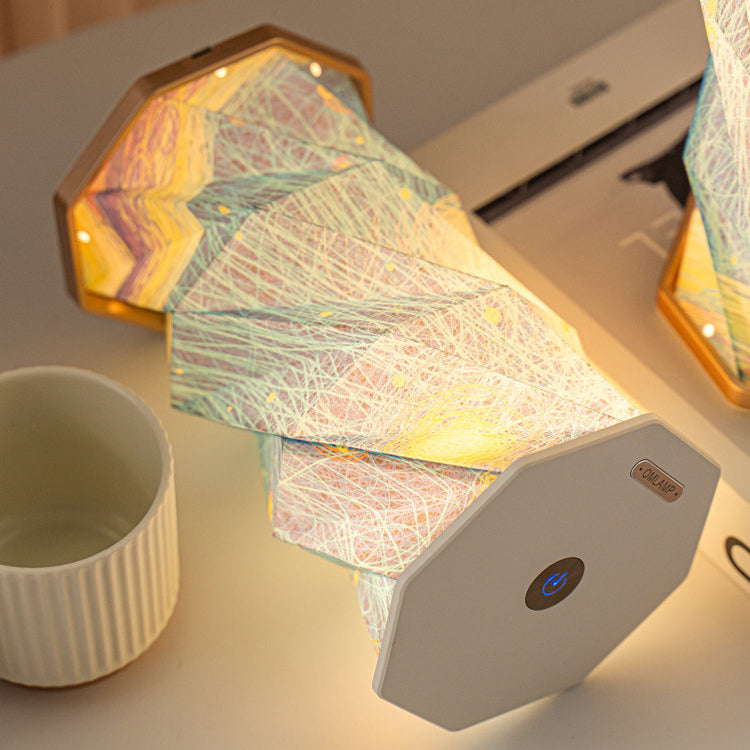 Starry Night folding lamp shown half-folded with one end closed and the other open, warm light spilling from the accordion paper shade printed with blue and gold starry swirls, another lamp fully lit in the background