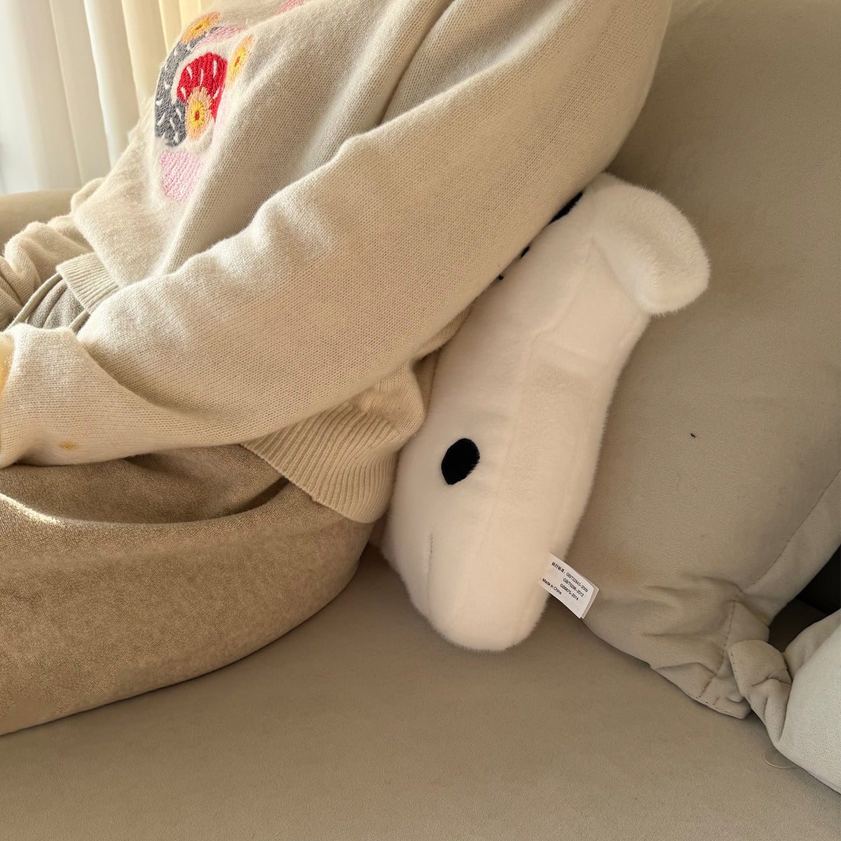 Close-up of the spotty white puppy hug pillow resting against a sofa cushion - silhouette body with black eye patch and dots, soft velvet pile, embroidered face and woven brand label on the side