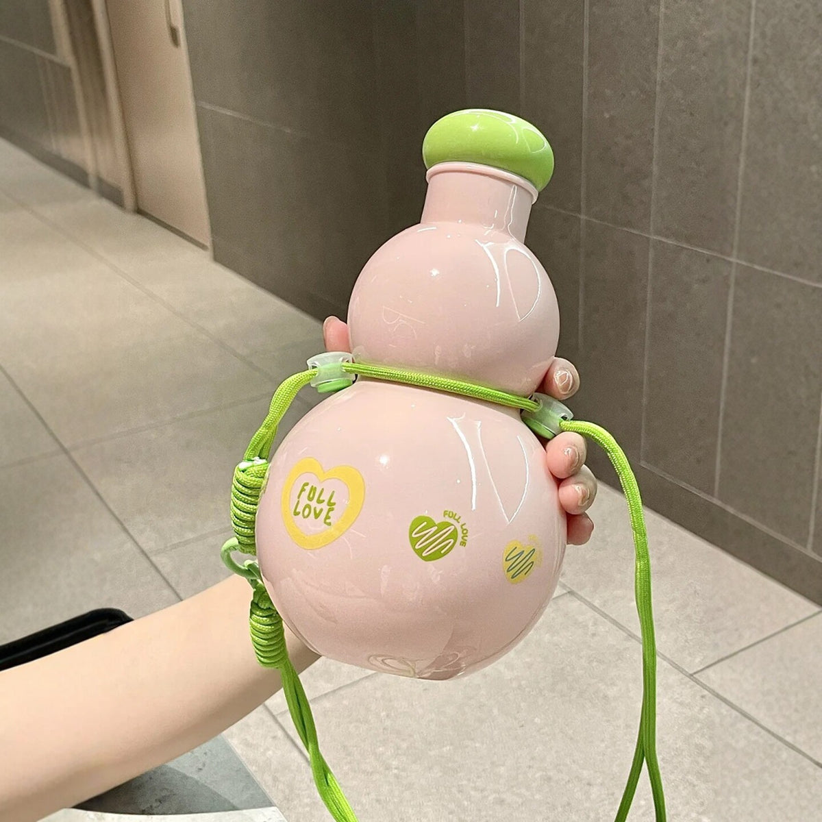 Hand holding the Sweet Pink gourd water bottle with its green lid on and green braided strap wound around the gourd waist - glossy pink body with FULL LOVE heart print in an indoor setting
