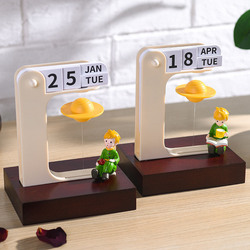 Two floating planet perpetual calendars side by side on a wooden desk - one with the rose prince showing 25 JAN TUE and one with the reading prince showing 18 APR TUE, both with golden Saturns floating in cream frames