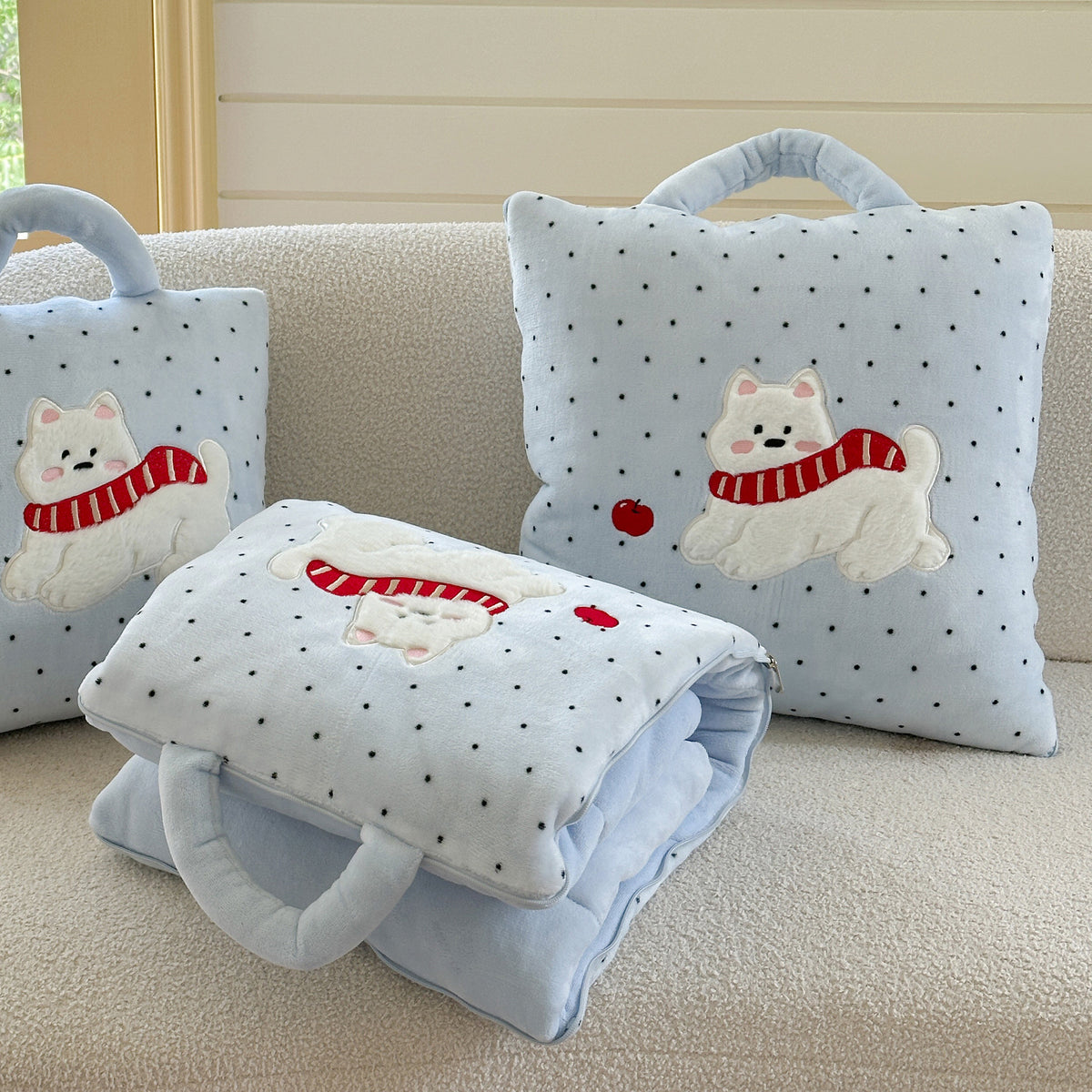 Puppy pillow blanket scene on a cream boucle sofa - unfolded baby blue polka dot blanket draped over the seat, two Westie cushions standing behind, pink keyboard and Christmas tree in the background, cozy winter living room