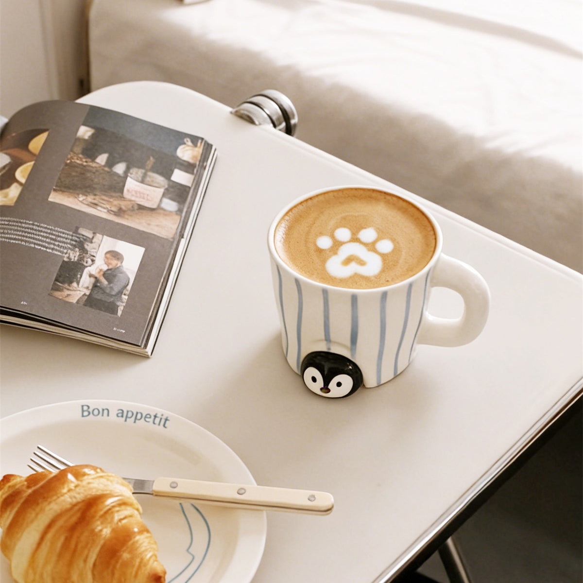 Peekaboo penguin mug with paw-print latte art on a white bedside table - glossy penguin peeking out from under the blue-striped ceramic cup, an open magazine and a Bon appetit plate with croissant nearby