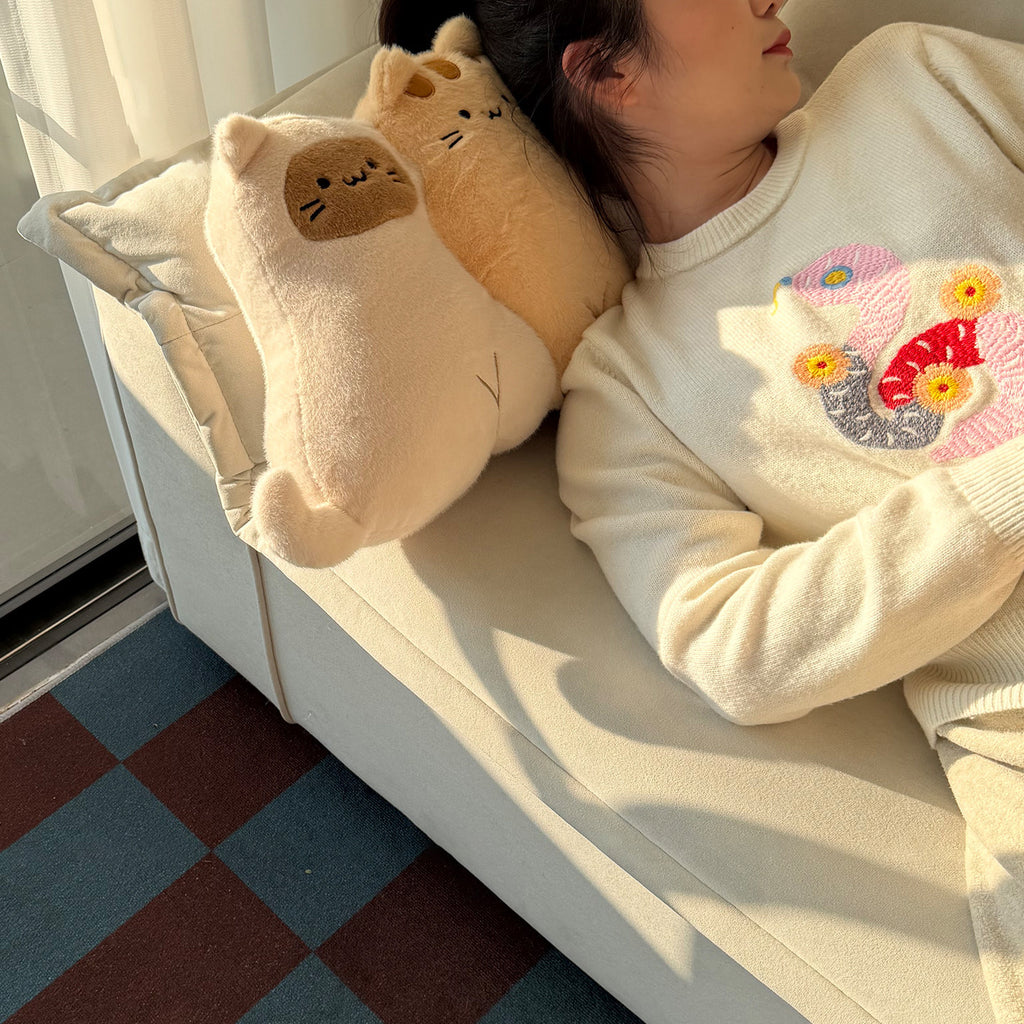 Woman napping on a sofa with two long-body cat hug pillows beside her head - cow-cat and ginger tabby plush cushions with embroidered sleepy faces, warm sunlight across the room