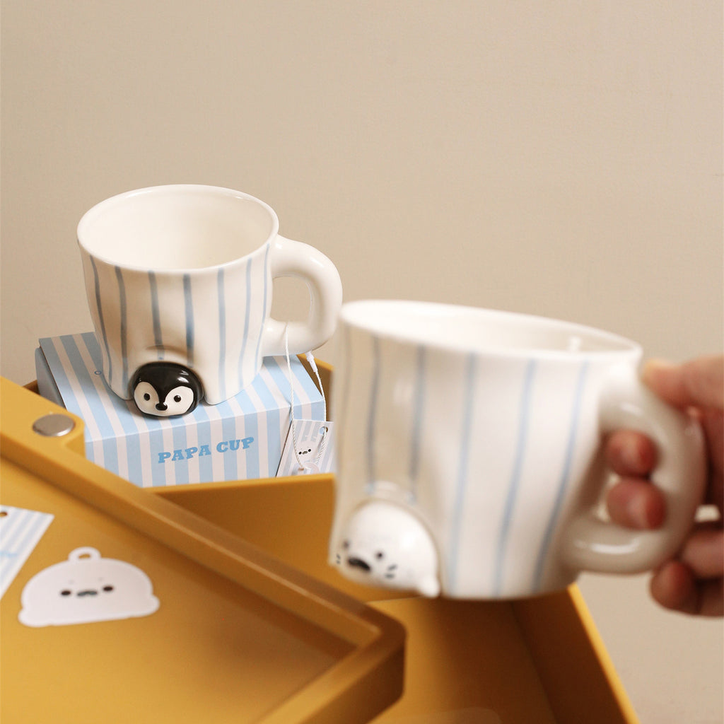 Peekaboo penguin mug sitting on its blue-striped gift box on a yellow tray, a hand holding the seal mug pouring in from the right - both hand-painted striped ceramic cups with animals peeking out, hang tags visible