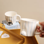 Peekaboo penguin mug sitting on its blue-striped gift box on a yellow tray, a hand holding the seal mug pouring in from the right - both hand-painted striped ceramic cups with animals peeking out, hang tags visible