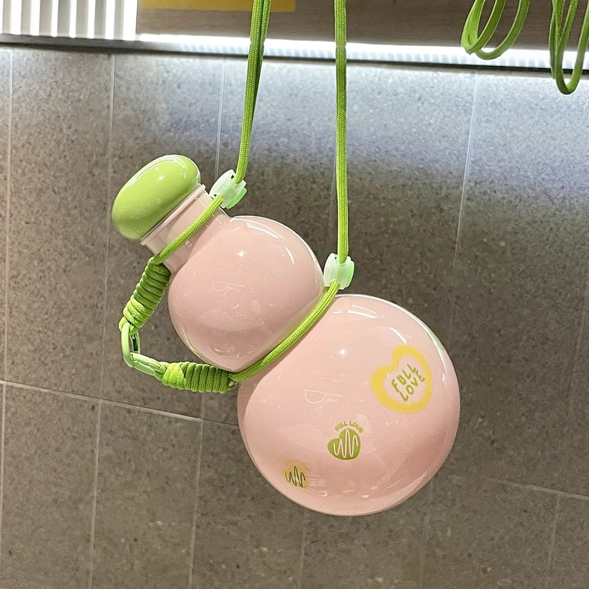 Sweet Pink gourd water bottle tied up by its green braided strap and hanging in the air - glossy pink gourd body with green lid and FULL LOVE prints, strap looped around the waist