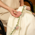 Close-up of a hand gently touching a hand-crocheted pink rose on the soft cream knit blanket.